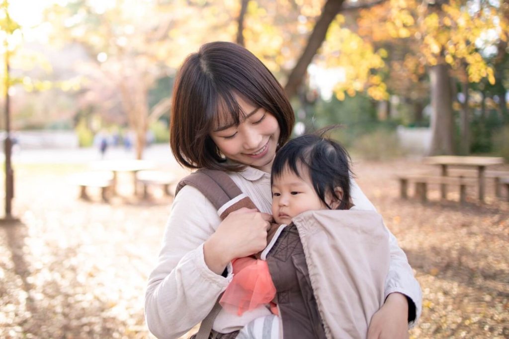 Maman souriante portant son bébé en porte-bébé dans un parc aux couleurs automnales dorées
