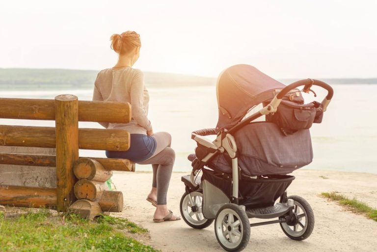Maman assise de dos sur un banc en bois face à un lac, la poussette de son bébé à ses côtés