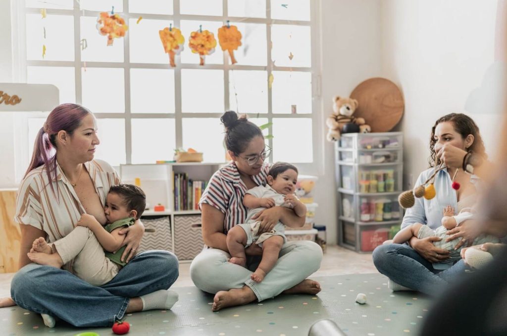 Trois mamans assises en tailleur sur un tapis à pois, chacune tenant son bébé, dans une salle lumineuse et chaleureuse