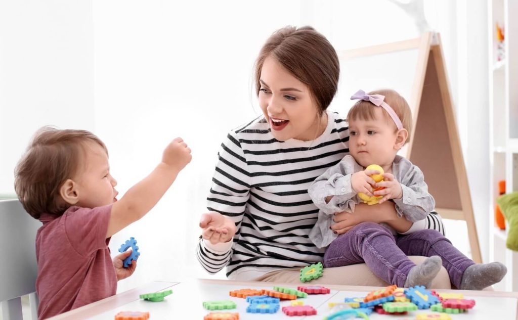 Une assistante maternelle souriante en pull rayé joue avec deux bébés assis à une table basse, entourés de jouets d'éveil colorés dans une pièce lumineuse.
