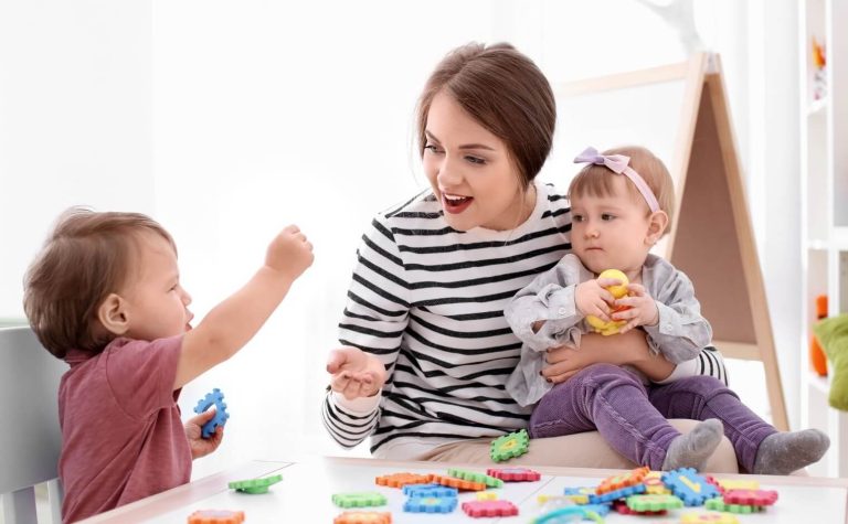 Une assistante maternelle souriante en pull rayé joue avec deux bébés assis à une table basse, entourés de jouets d'éveil colorés dans une pièce lumineuse.