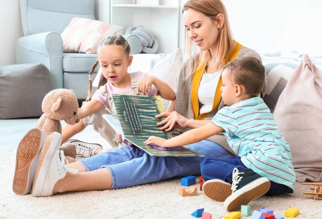 Une assistante maternelle assise sur un tapis lit un livre illustré à deux enfants en bas âge dans un salon chaleureux, avec des cubes colorés et un doudou autour d'eux.