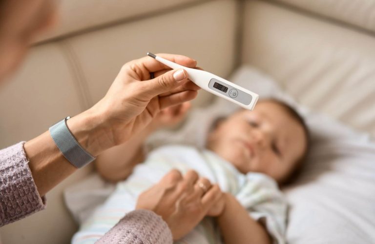 Une main tient un thermomètre numérique devant un bébé allongé sur un canapé, illustrant la gestion de l'enfant malade lors de la garde.