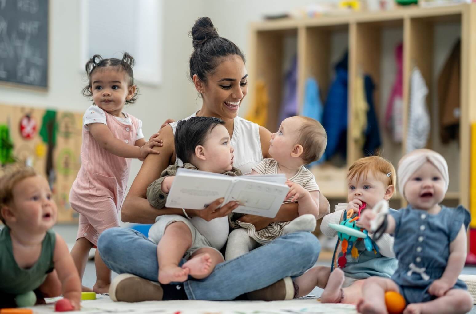Une professionnelle de la petite enfance souriante est assise en tailleur au milieu d'un groupe de six bébés dans une salle de crèche moderne, avec des casiers en bois et du matériel pédagogique en arrière-plan.