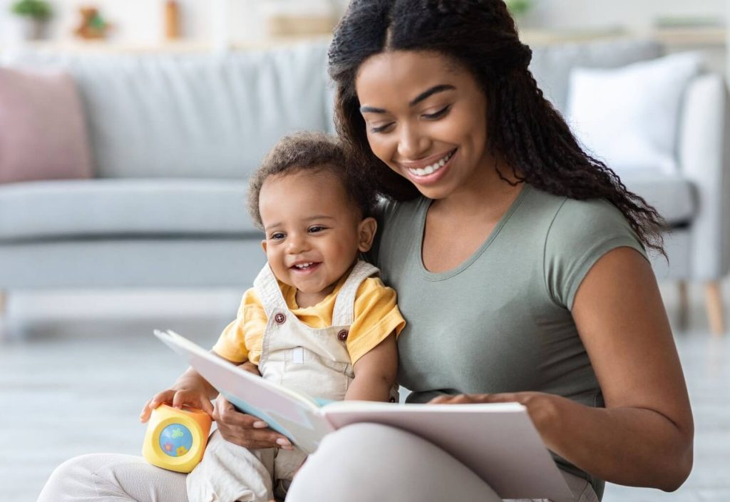 Une jeune femme souriante assise au sol tient un bébé heureux sur ses genoux et lui lit un livre illustré dans un intérieur lumineux et épuré.
