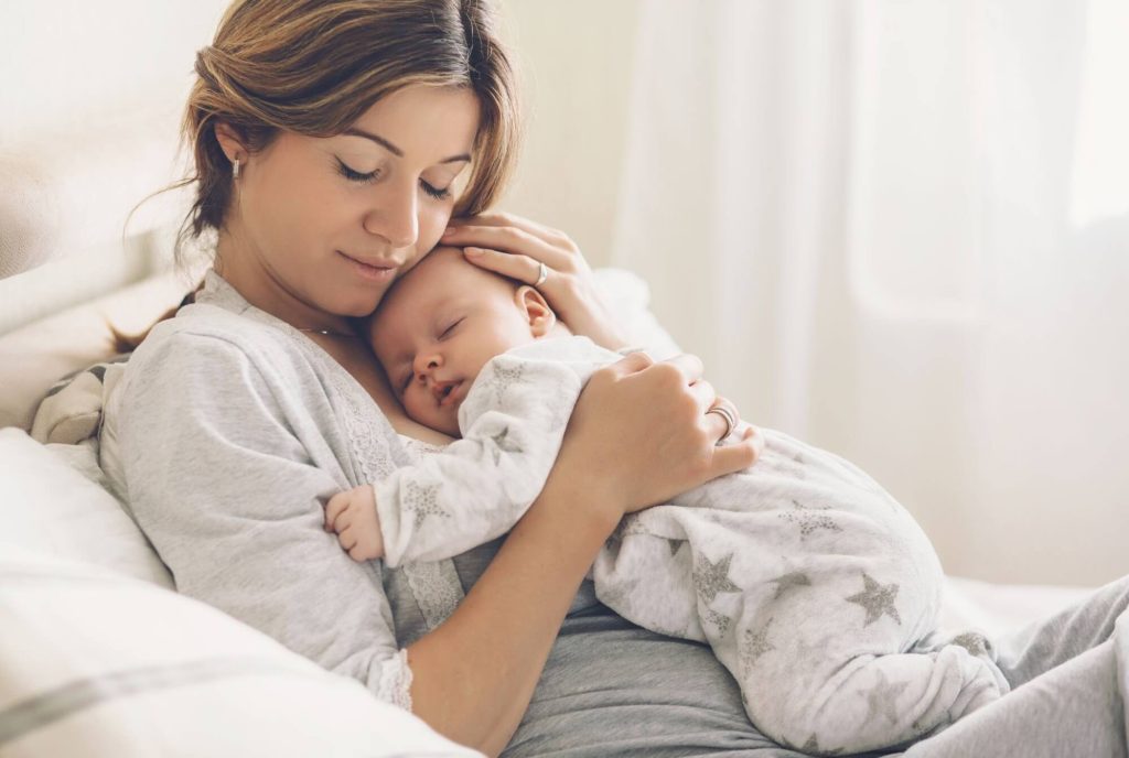 Jeune maman en pyjama tenant son nouveau-né endormi contre elle — livres post-partum