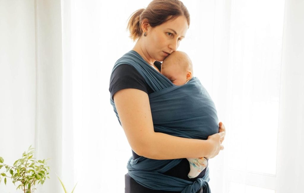 Maman portant son nouveau-né dans une écharpe élastique bleu pétrole à la maison, moment de tendresse et de sérénité
