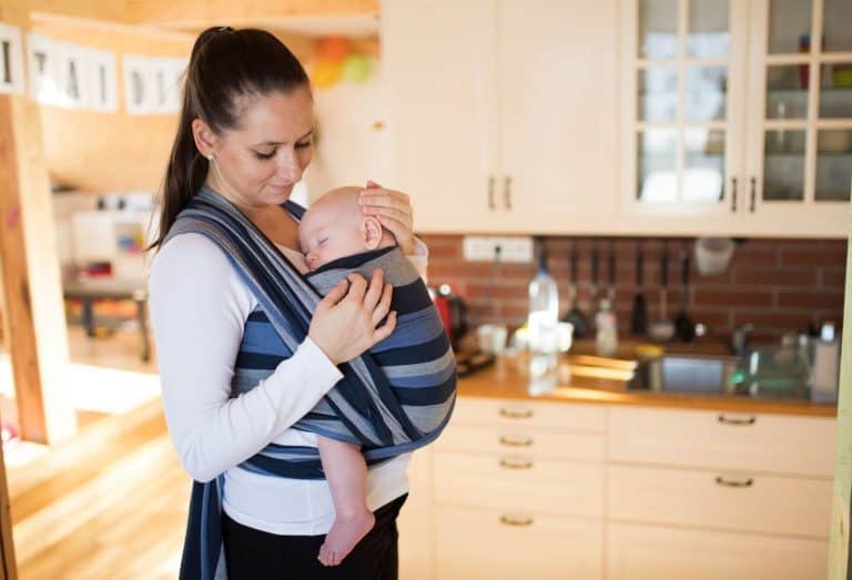 Parent portant son bébé en écharpe tissée rayée à la maison dans la cuisine, illustrant le portage au quotidien avec les mains libres