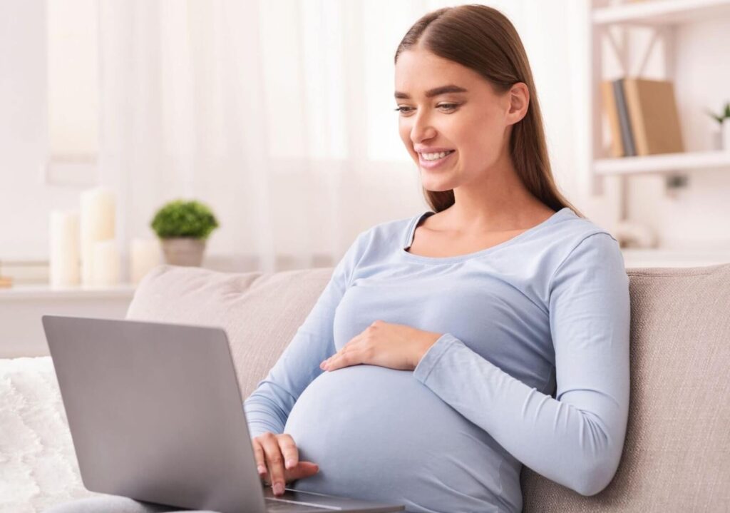 Femme enceinte souriante sur un canapé consultant ses démarches administratives sur un ordinateur