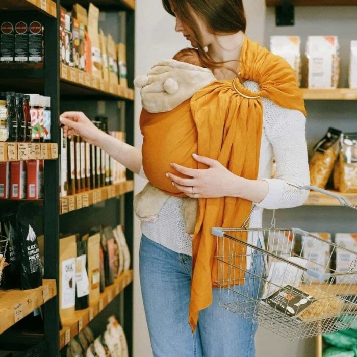 Parent portant son nouveau-né dans une sling orange tout en faisant les courses, démontrant la praticité et les mains libres